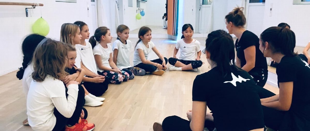 Kinder und Trainerinnen sitzen im großen Kreis auf dem Studioboden